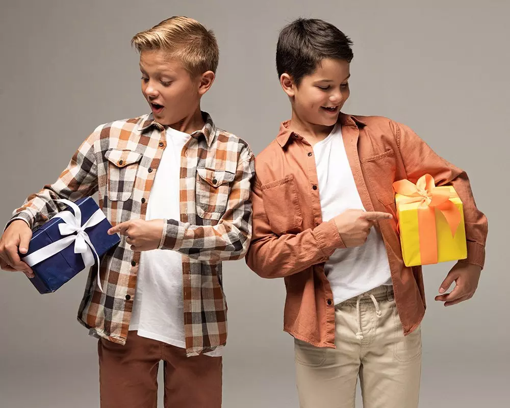 Dwóch chłopców w koszulach Pepco, jeden w kraciastej, drugi w pomarańczowej, trzymających kolorowe prezenty z wstążkami.