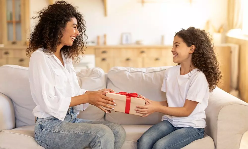 Žena a dívka v bílých tričkách a džínách od Pepco si předávají dárkovou krabici s červenou mašlí na pohovce v útulném domově.