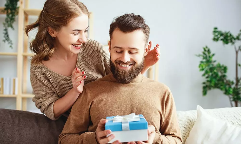 Para uśmiecha się podczas otwierania niebieskiego prezentu z białą wstążką. Oboje noszą swetry w kolorze beżowym od Pepco.