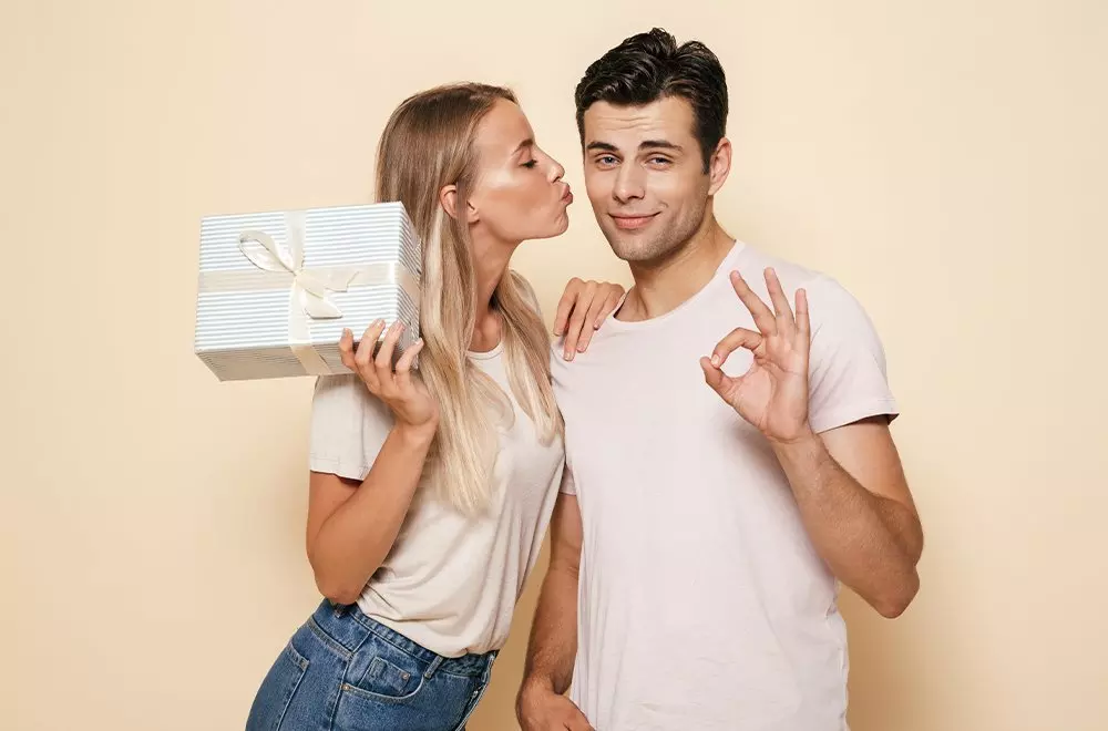 Para trzymająca ozdobne pudełko prezentowe od Pepco, ubrana w białe koszulki, pokazująca gest OK na beżowym tle.