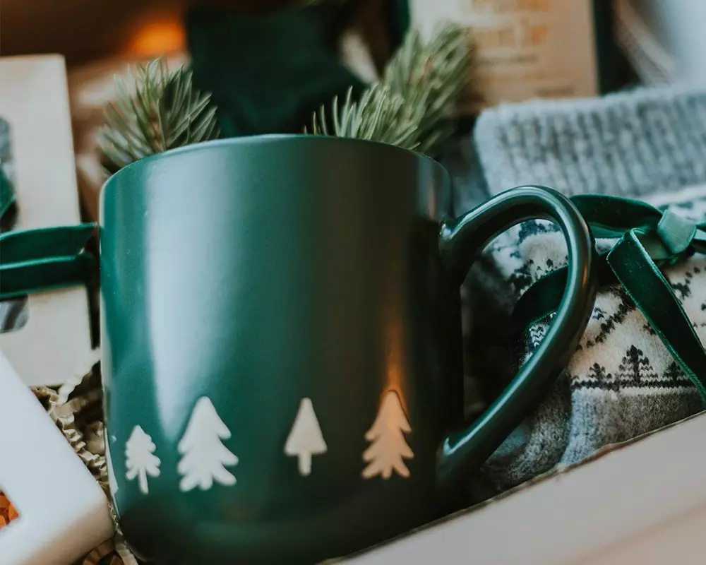 Zielony kubek Pepco z motywem choinek, ozdobiony gałązkami świerku, na zimowym tle z dzianinowym kocem.