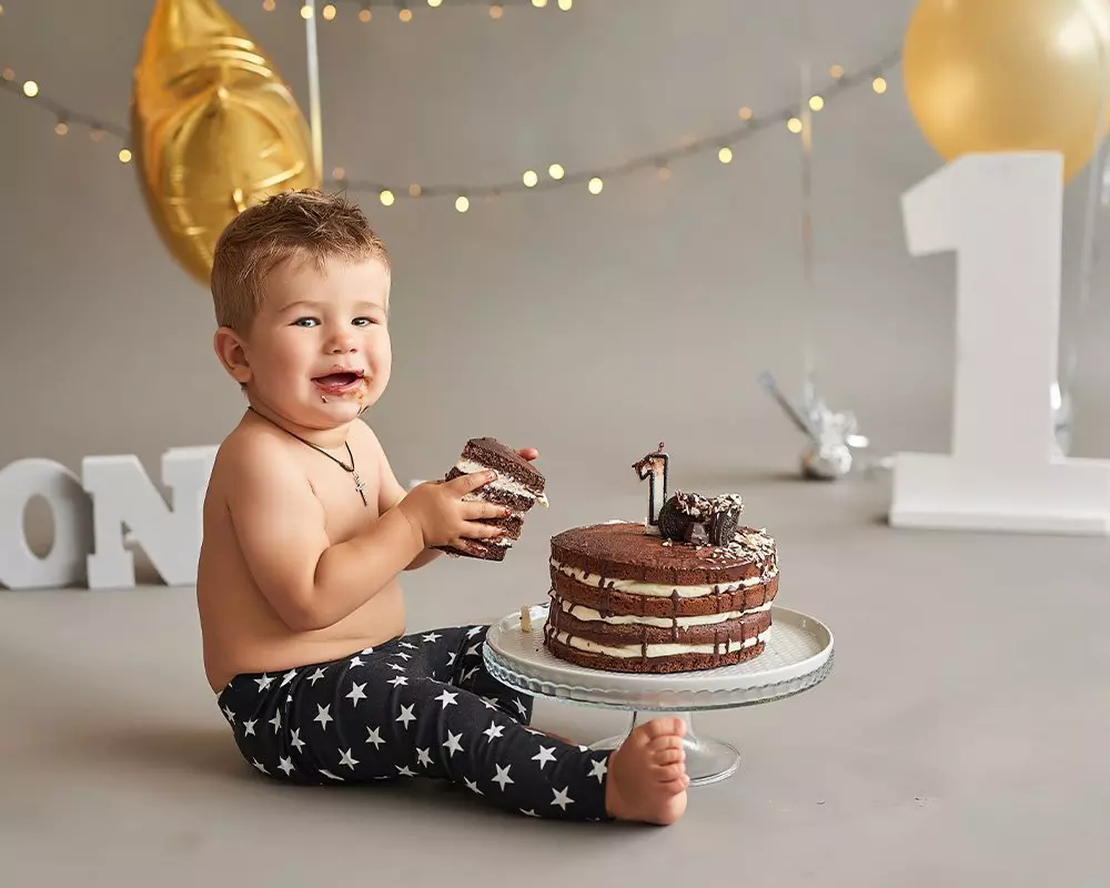 Child enjoying first birthday celebration with chocolate cake, wearing Pepco star-patterned pants amid festive decorations.