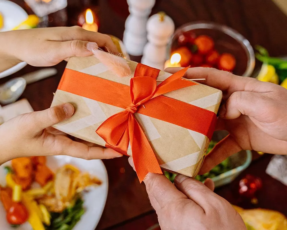 Kezek átnyújtanak egy narancssárga masnival díszített Pepco ajándékot egy ünnepi asztalnál, gyertyákkal és étellel körülvéve.