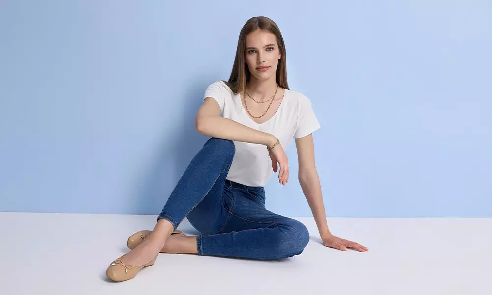 Woman sitting wearing Pepco white t-shirt, blue jeans and gold necklaces with beige ballet flats.