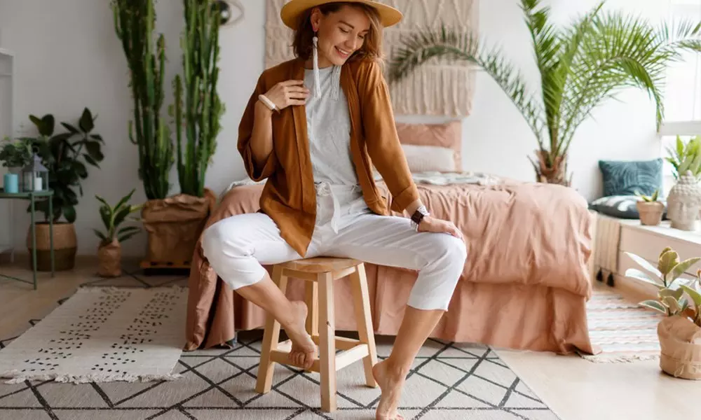 Osoba w brązowej marynarce i białych spodniach od Pepco siedząca na drewnianym stołku w przytulnej sypialni z roślinami.