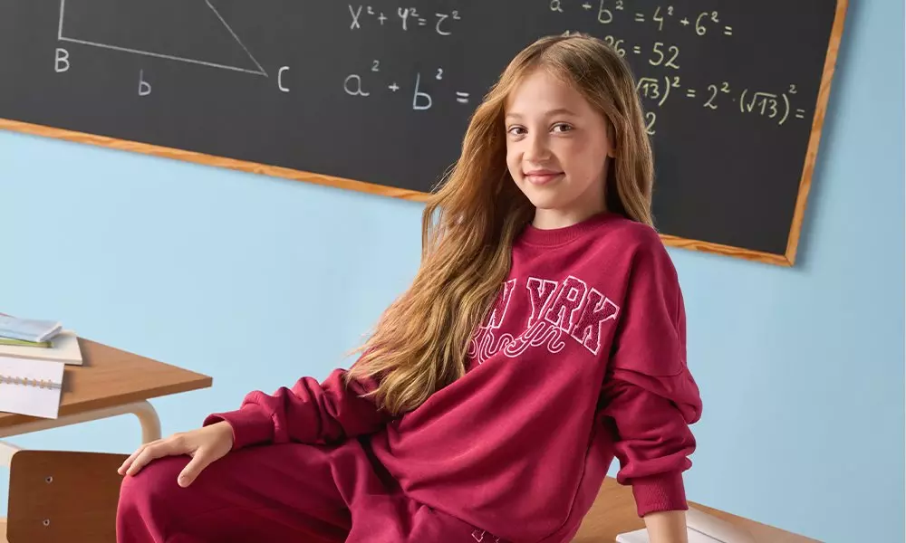 Girl wearing Pepco burgundy sweatshirt with text detail sitting at desk in classroom with maths equations on blackboard.