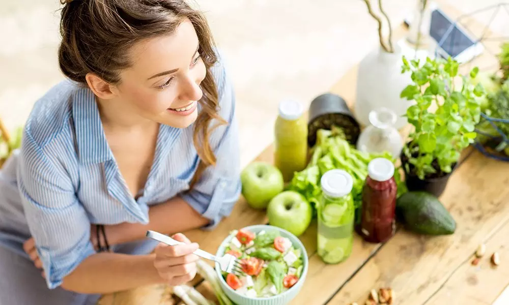 Osoba w niebieskiej koszuli jedząca sałatkę otoczona świeżymi owocami, warzywami i ziołami z Pepco na drewnianym stole.