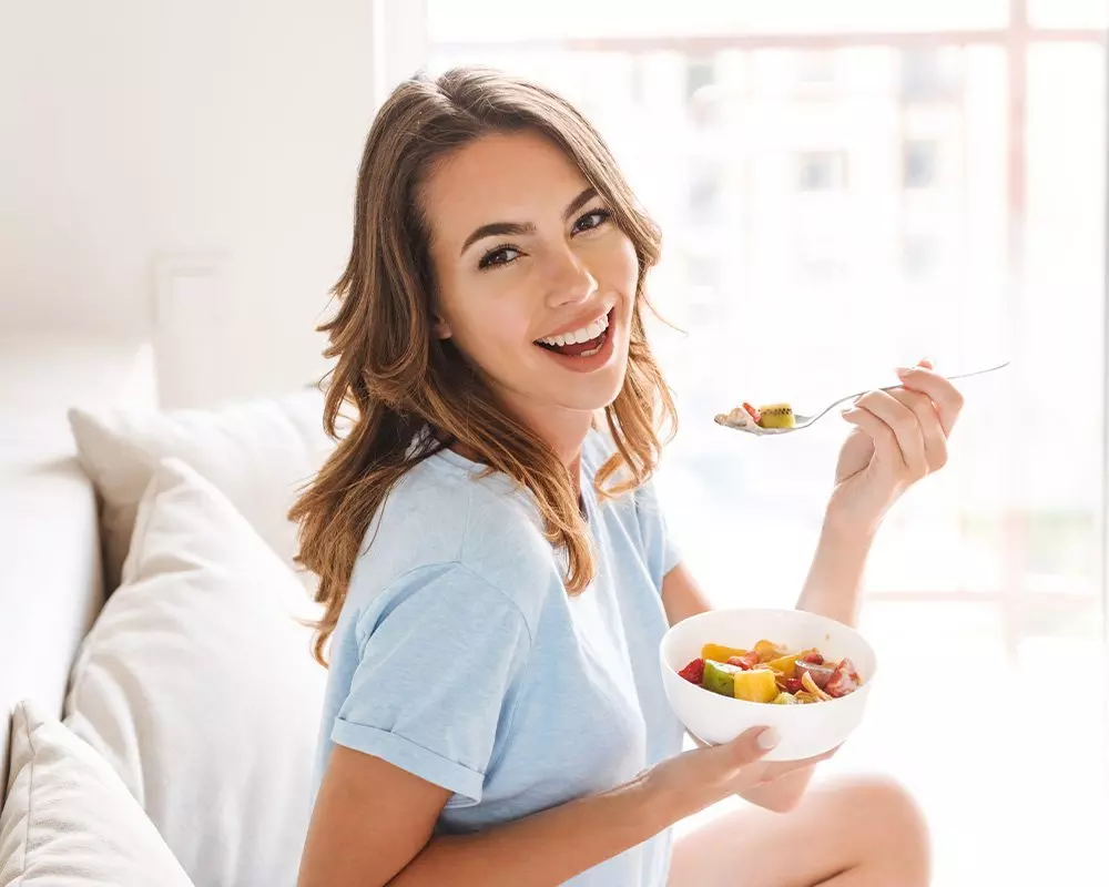 Kobieta w niebieskiej koszulce Pepco uśmiecha się jedząc sałatkę owocową z białej miski w jasnym, przytulnym wnętrzu.