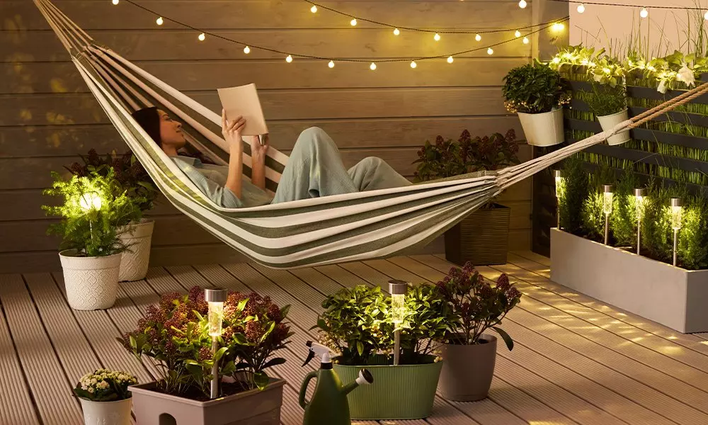 Person reading in striped hammock on wooden deck surrounded by potted plants and string lights.
