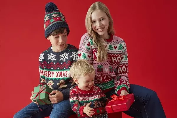 Famiglia Pepco che indossa maglioni natalizi coordinati e tiene regali su sfondo rosso.