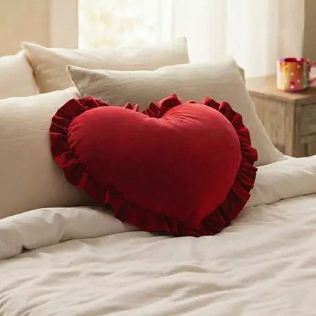 Czerwona poduszka w kształcie serca z falbanką od Pepco na jasnej pościeli, obok kolorowy kubek na stoliku.