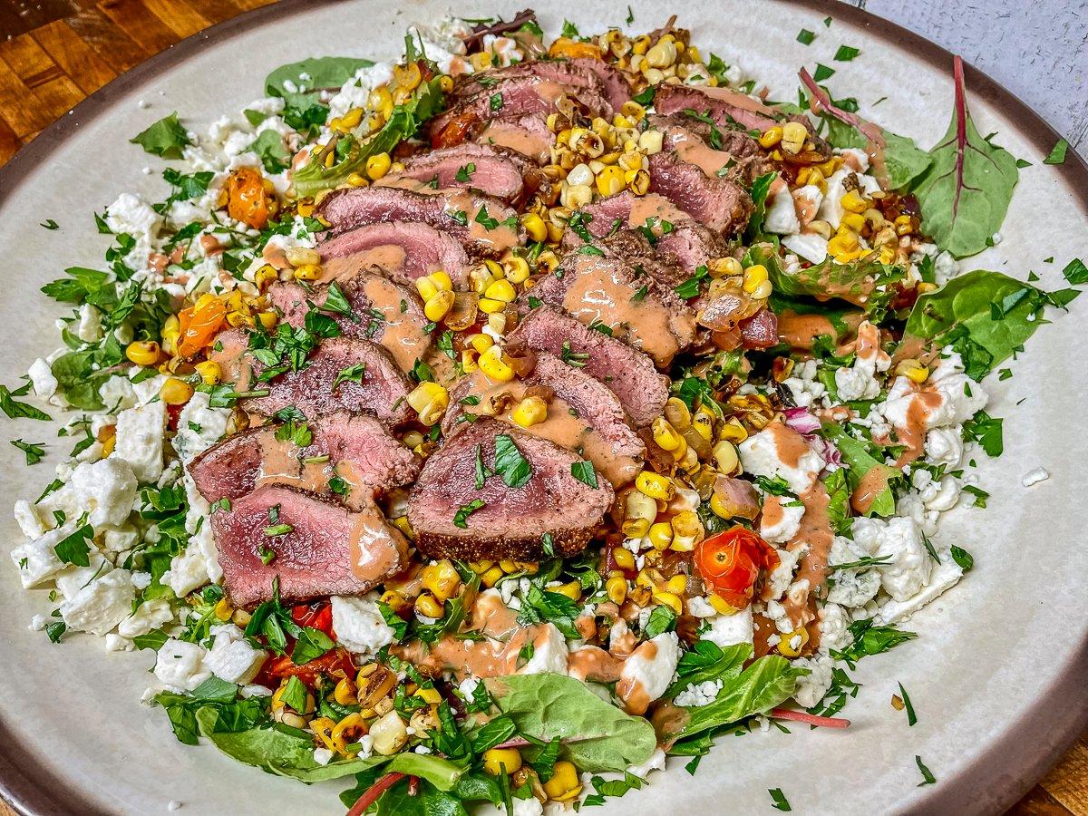 Layers of grilled sweet corn, red onion, and fresh tomatoes get layered over crisp salad greens and topped with thinly sliced backstrap.