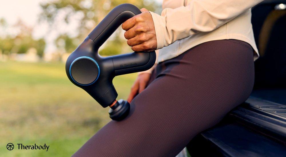 a woman massaging her leg with a theragun leaning on a car