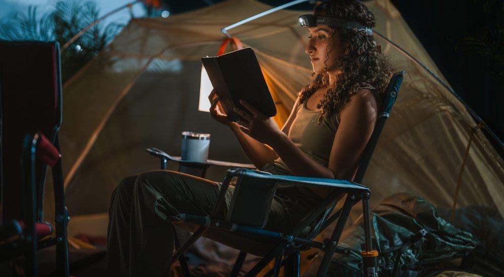 a girl reading a book with a headlamp on at a campsite