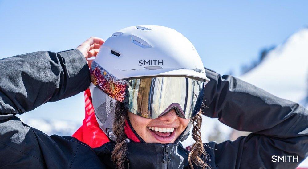 a girl strapping on her ski googles on the top of a mountain