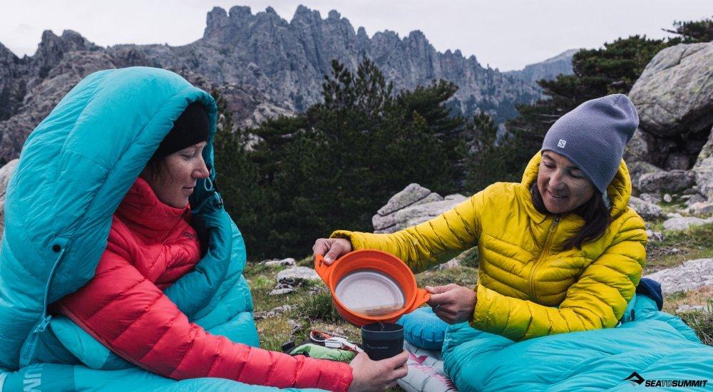 two women wrapped up in a sleeping bag in the outdoors while pouring coffee