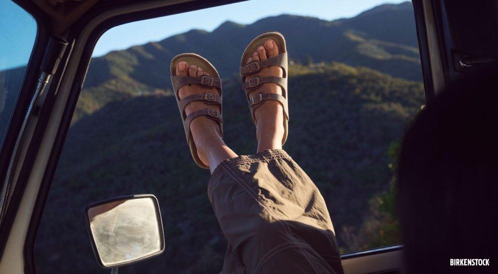 A woman leans back in the seat of a car with her feet out the window wearing BIRKENSTOCK sandals