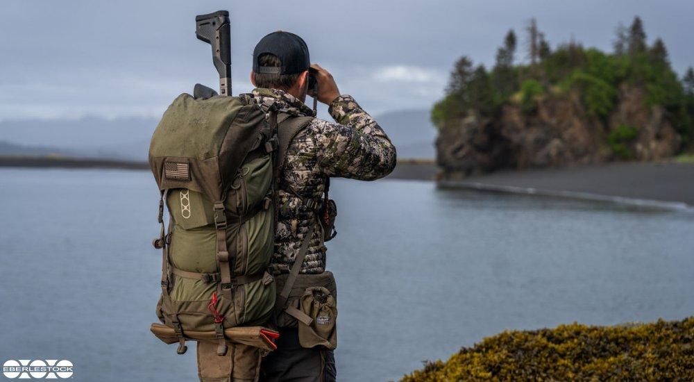 a man wearing a hunting backpack looking through his binoculars looking at an island