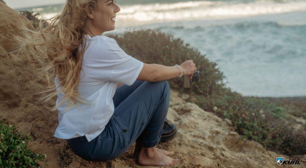 A woman sits overlooking the ocean wearing Kuhl pants