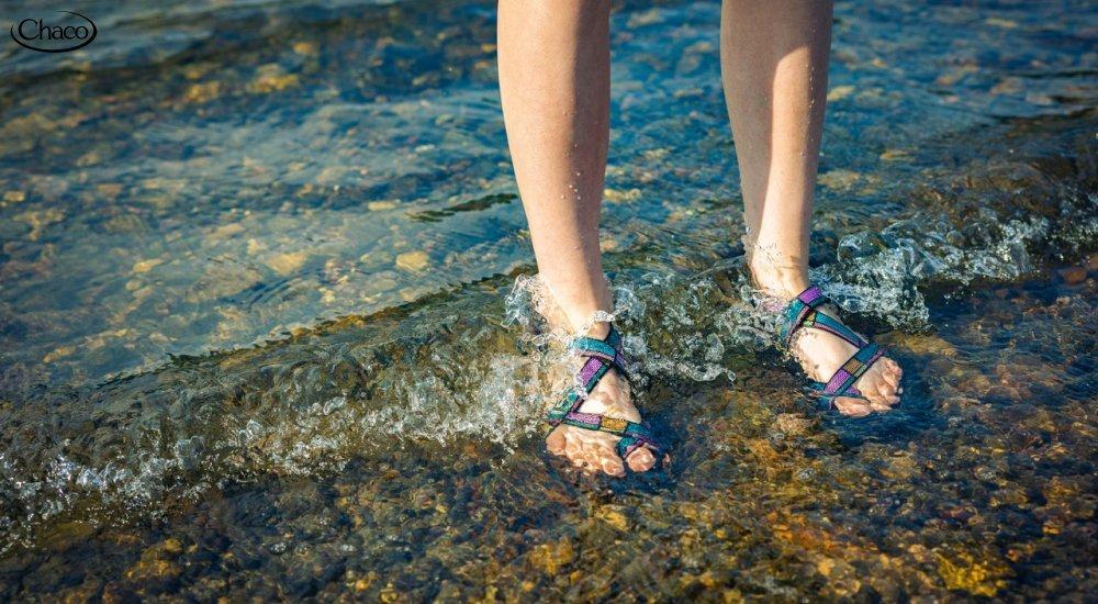 A woman stands with her feet submerged in a lake wearing Chacos 