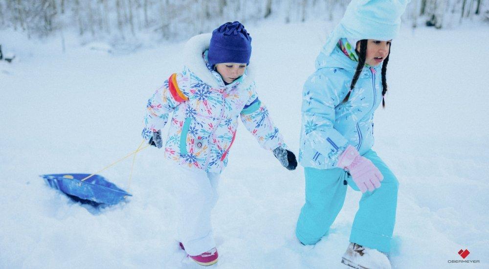 Two girls trudge through snow in winter gear while one pulls a sled