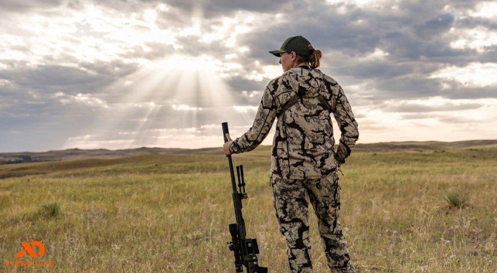 a woman standing in the field with a gun and the sun peeking through the clouds