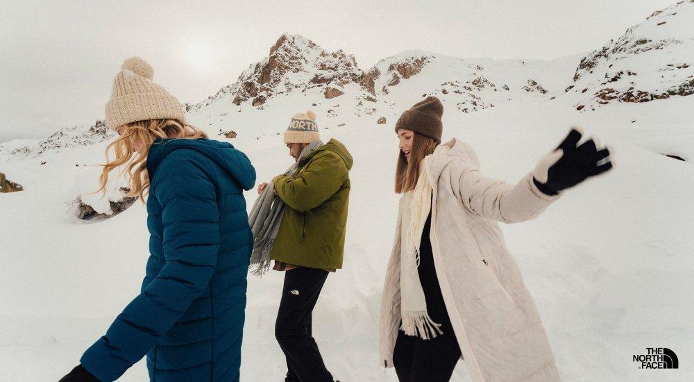 Three women wearing hats and puffer jackets in a snowy mountain scene