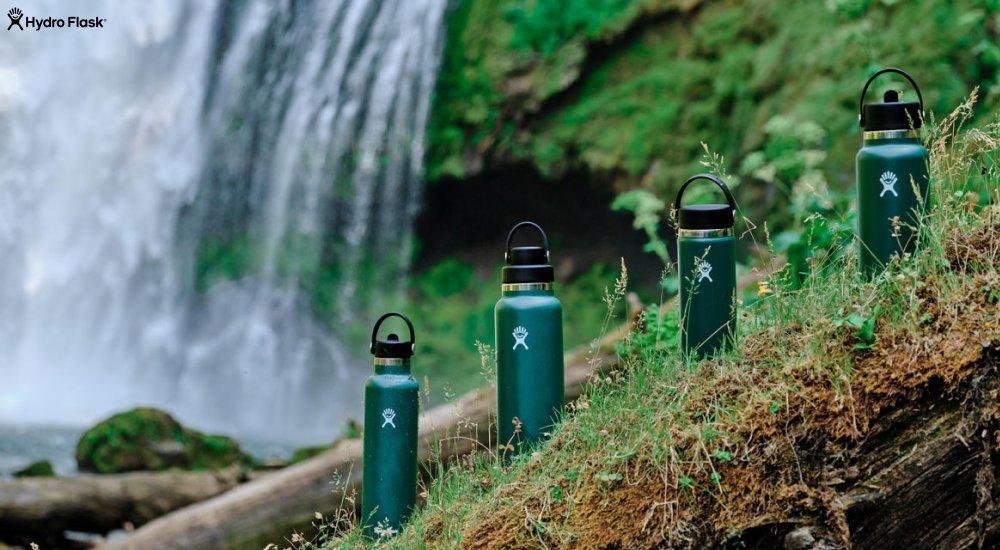 four green hydroflask bottle lined up in the rain forest next to a waterfall