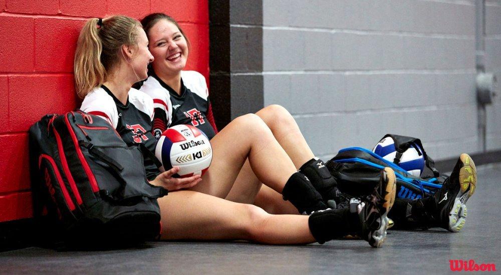 two girls sitting against the gym wall with their volleyballs and volleyball bags