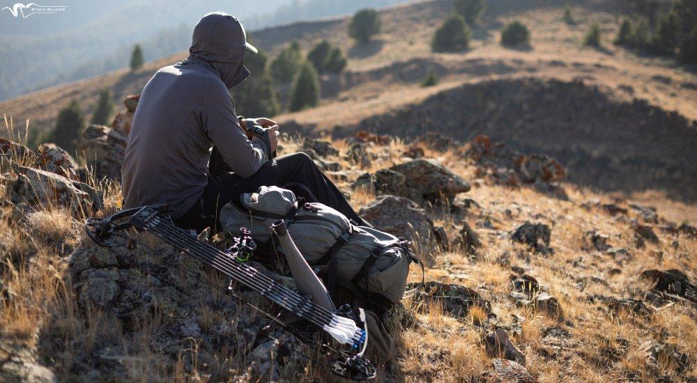 an elk hunter sitting on a bluff with his hunting pack and bow next to him