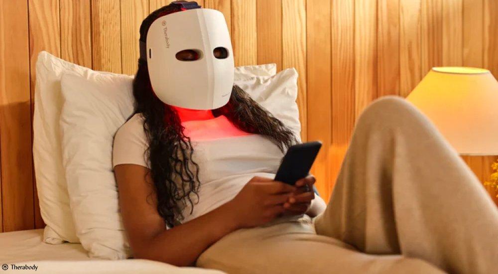 a woman laying in her bed with a light therapy mask on while she looks at her phone