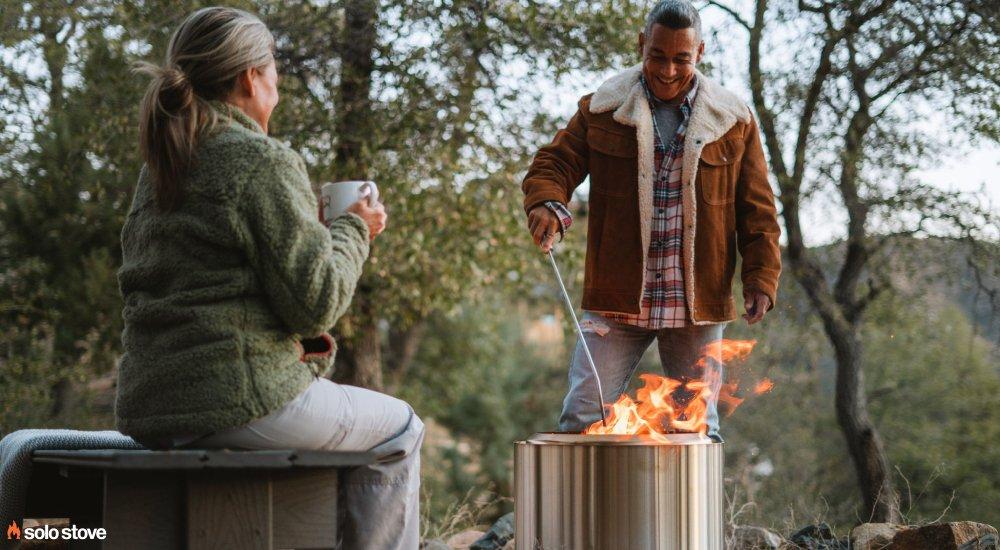 a couple sitting by a solo stove in the woods while drinking coffee
