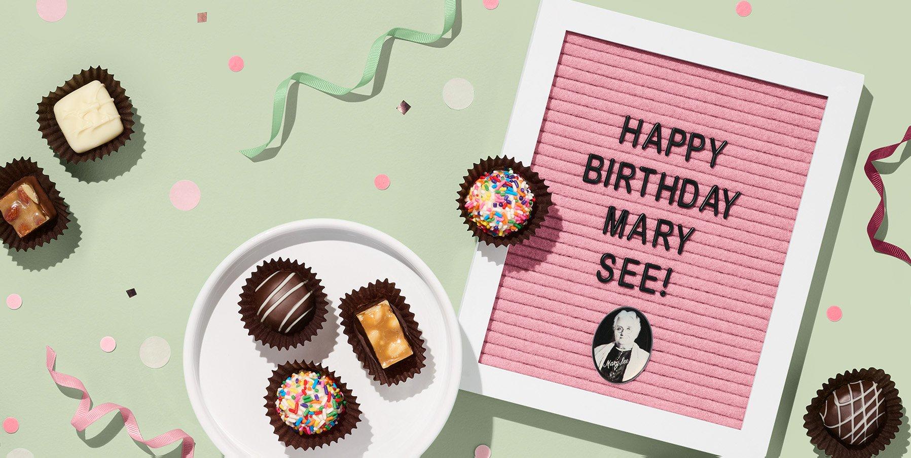 A letter board with "Happy Birthday Mary See!" beside a photo of a woman and surrounded by various chocolates in paper cups, on a green background with confetti and ribbons.
