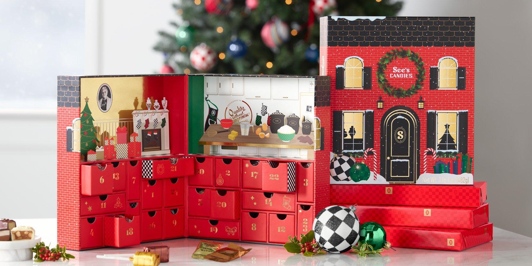 An advent calendar from See's Candies is displayed with its compartments open, revealing small drawers. The exterior resembles a decorated house with a wreath and festive ornaments. In the background, there is a Christmas tree adorned with lights and baubles.