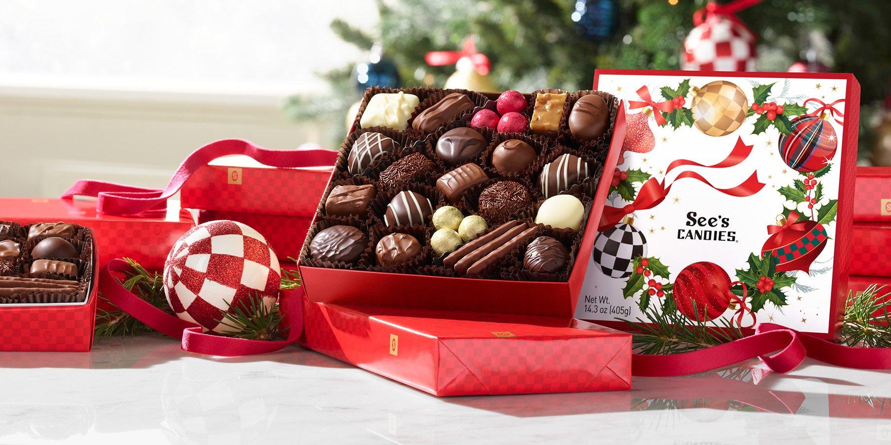 A box of chocolates with a design of a festive wreath made of ornaments sits in front of a christmas tree