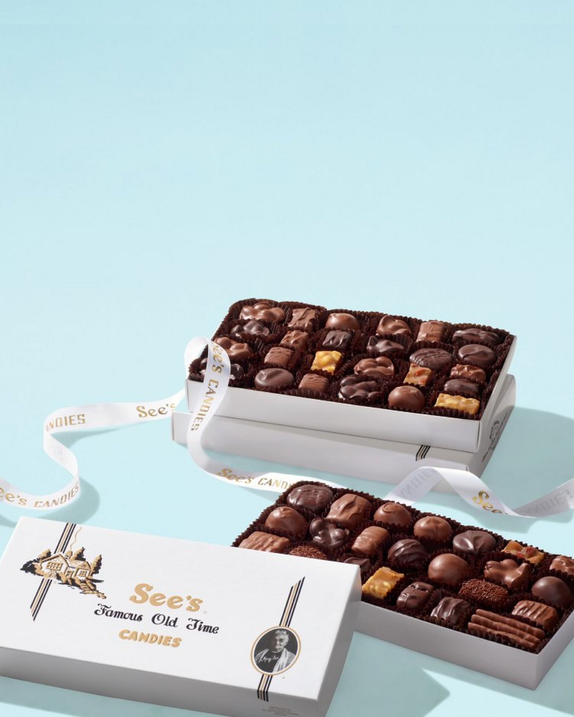 Open boxes of See's Candies assorted chocolates on a light blue background.