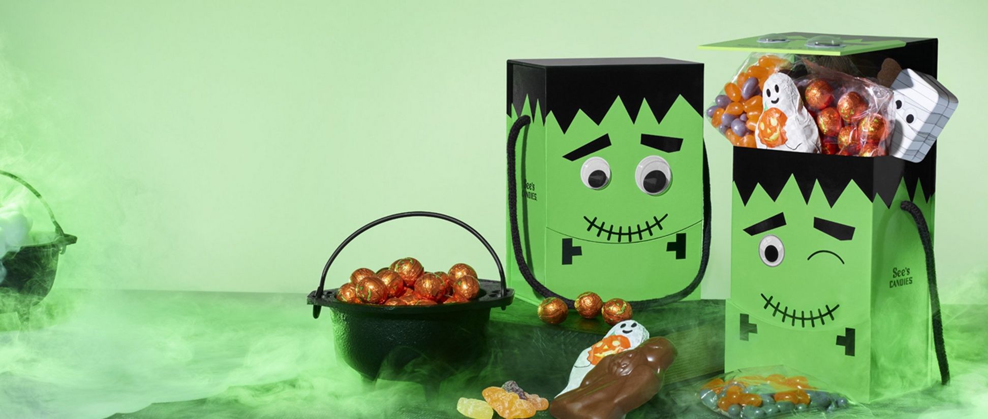 TwoFrankenstein shaped candy boxed with googly eyes are shown with candy spilling out of them. There is green smoke surrounding the image.