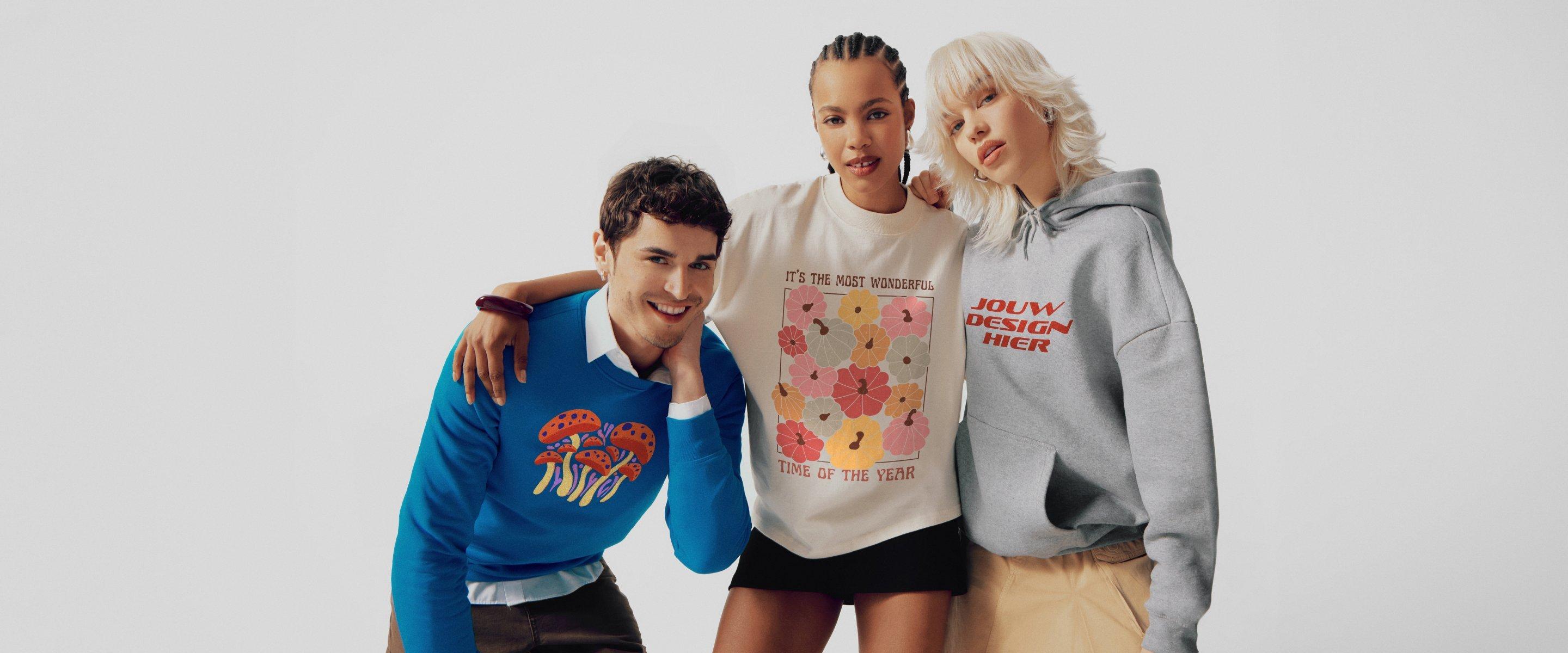 Drie modellen poseren samen: Man in een blauwe trui met paddenstoelenmotief, vrouw in een crèmekleurige trui met bloemenmotief, vrouw in een grijze hoodie met rode letters.