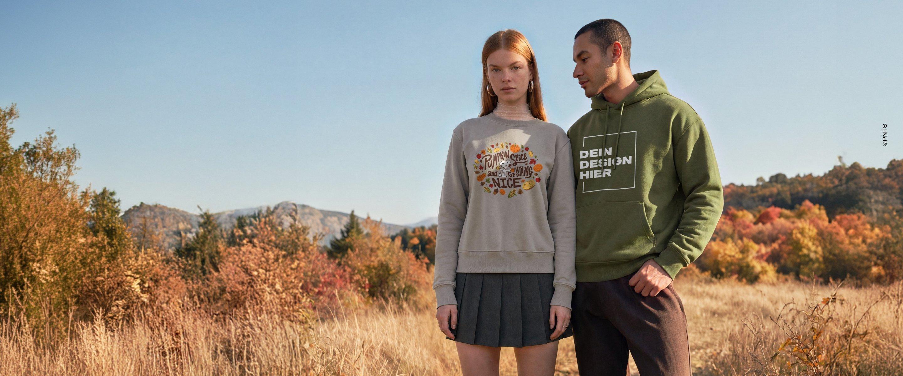 Frau im grauen Sweatshirt mit herbstlichem Kürbismotiv und Mann im grünen Hoodie mit Schriftzug „Dein Design hier“ stehen vor herbstlicher Landschaft.