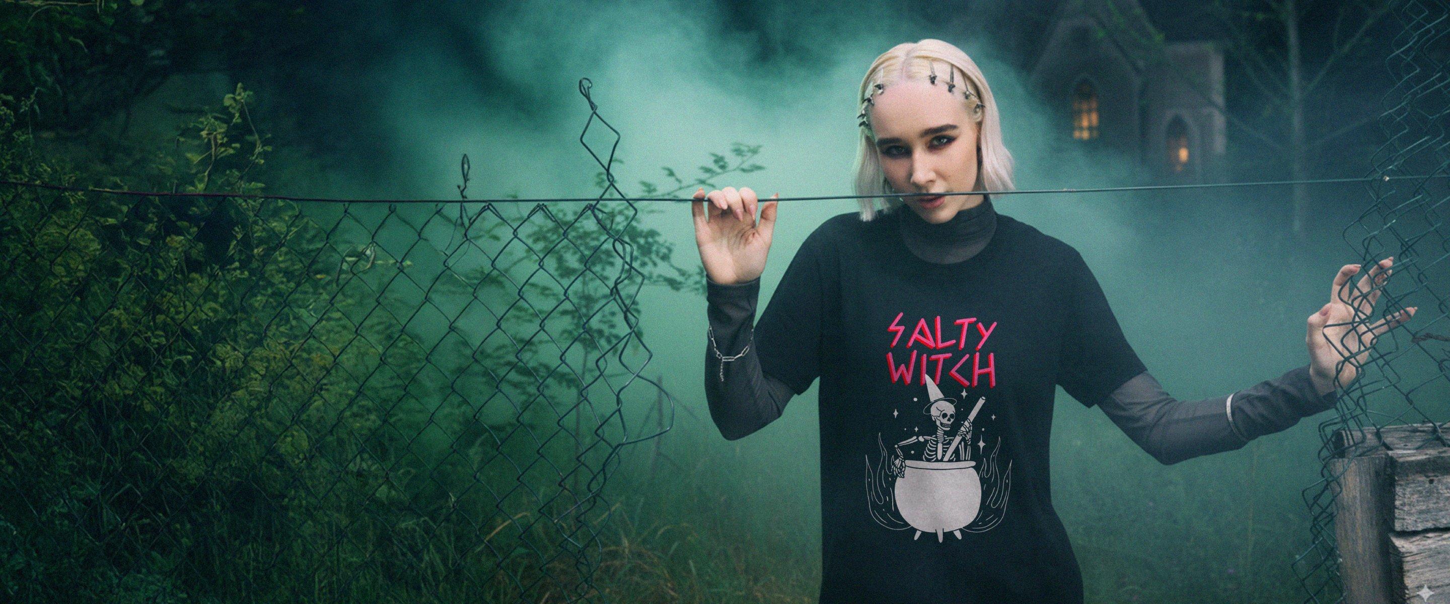 Vrouw met blond haar, gekleed in een zwart T-shirt met 'Salty Witch'-opdruk, staand voor een mistige achtergrond.