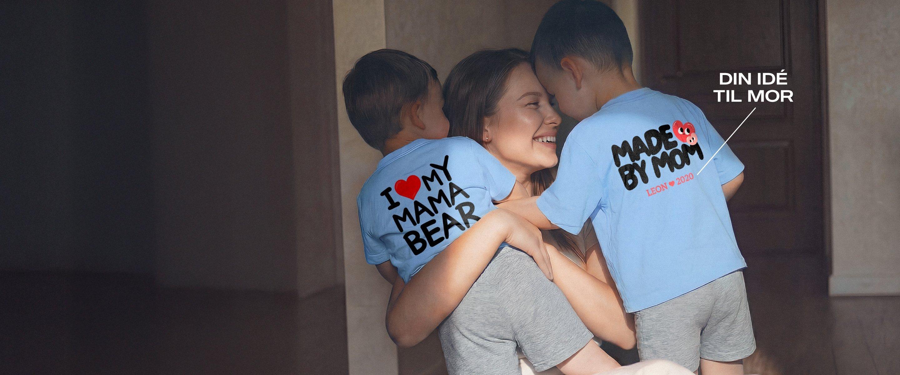 Smilende mor holder to små børn i armene; begge bærer blå T-shirts med mors dags-motiver og personlige tryk.