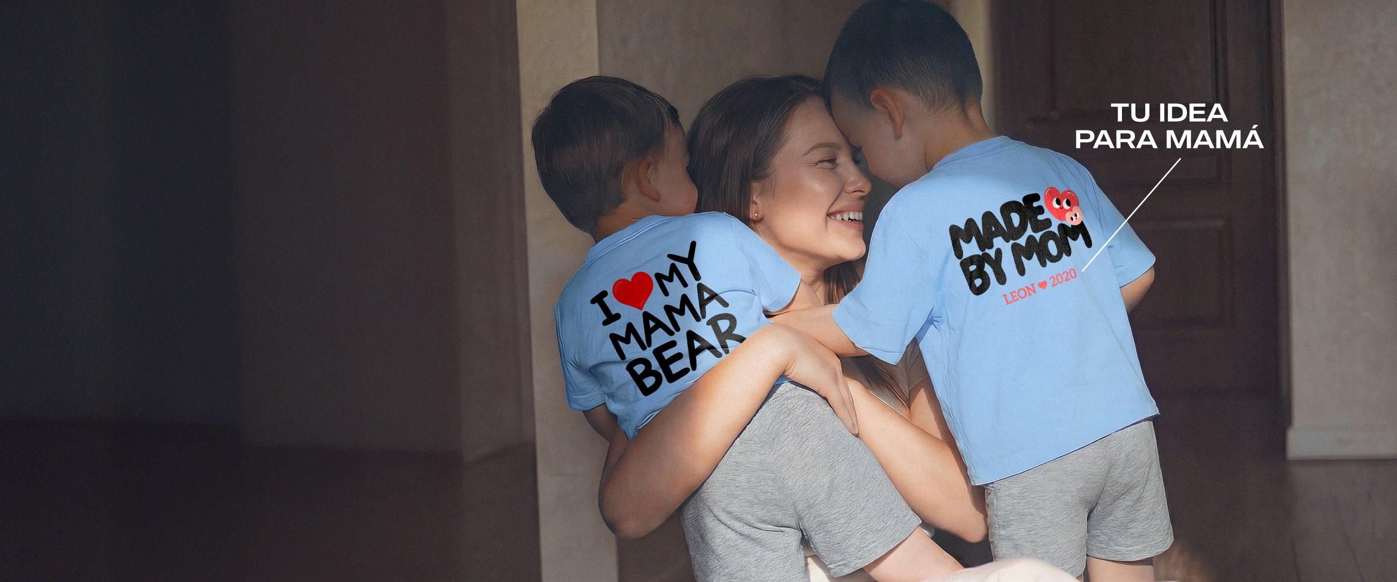 Madre sonriente sostiene a dos niños pequeños en brazos; ambos llevan camisetas azules con motivos del Día de la Madre y estampados personalizados.