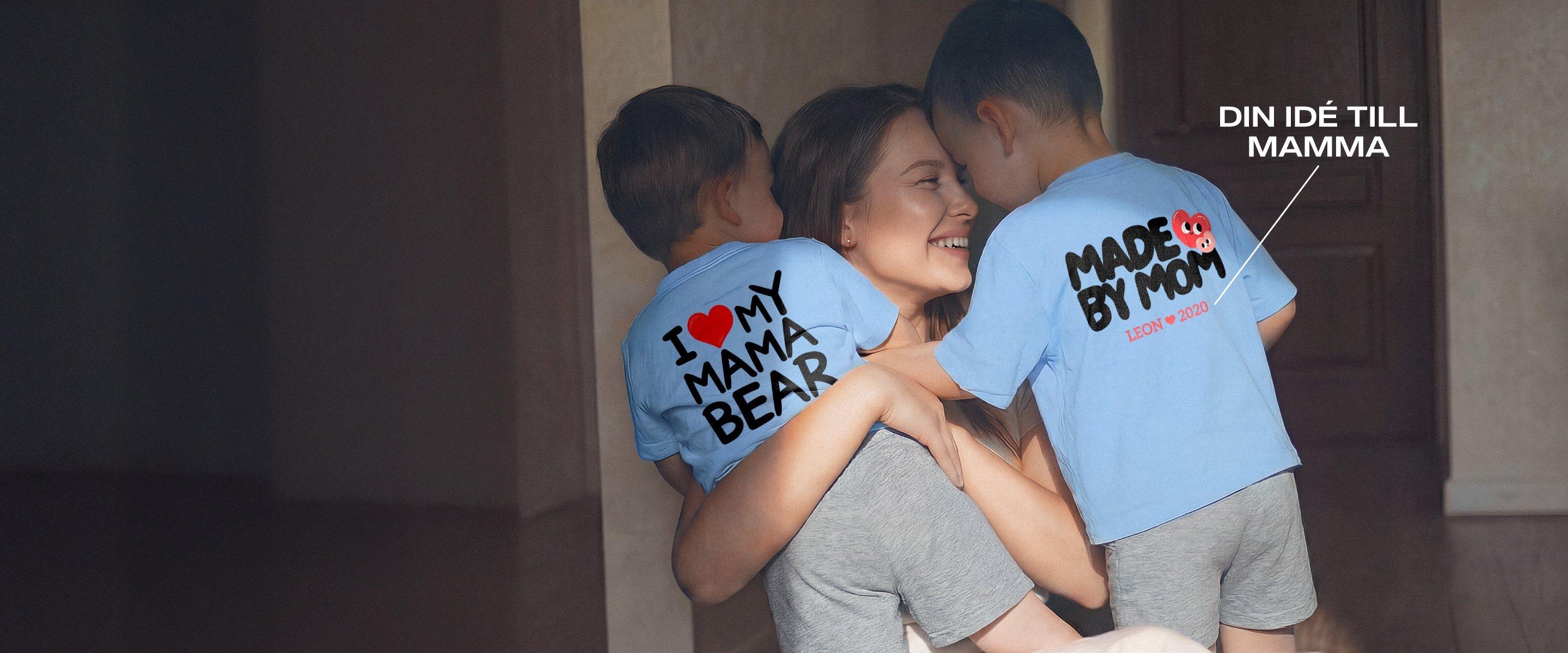 Leende mamma håller två små barn i famnen; båda bär blå T-shirts med mors dags-motiv och personliga tryck.