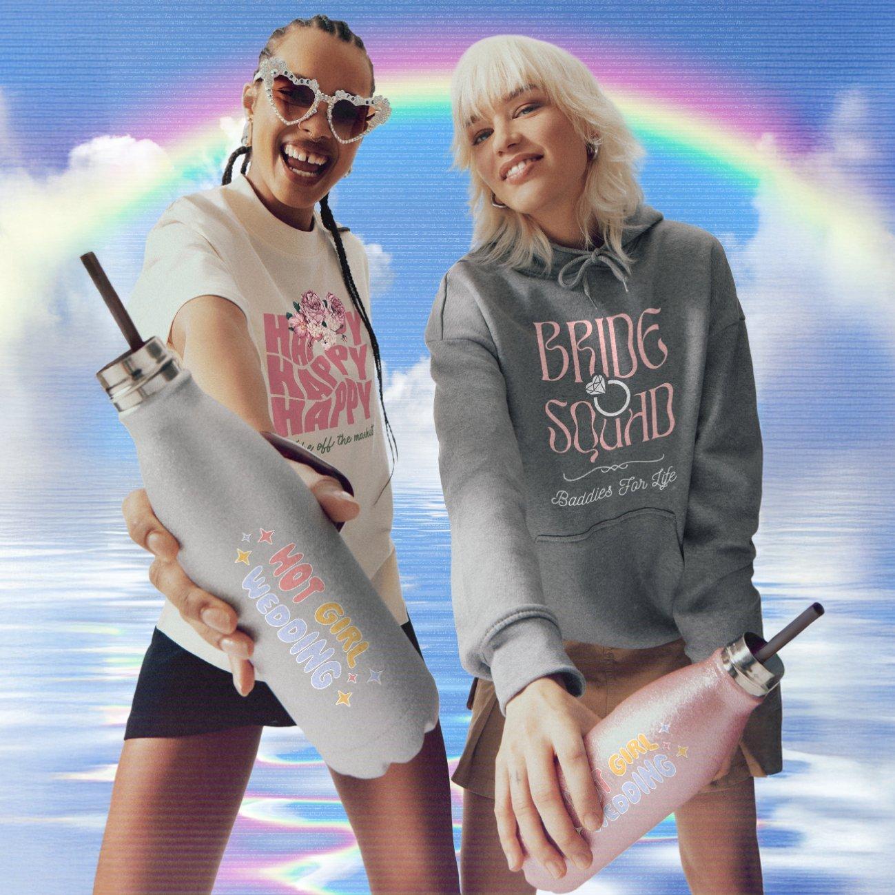 Dos mujeres frente a un fondo azul claro. Una lleva gafas de sol en forma de corazón y una camiseta blanca estampada, y sostiene una botella. La otra lleva una sudadera gris con letras rosas.