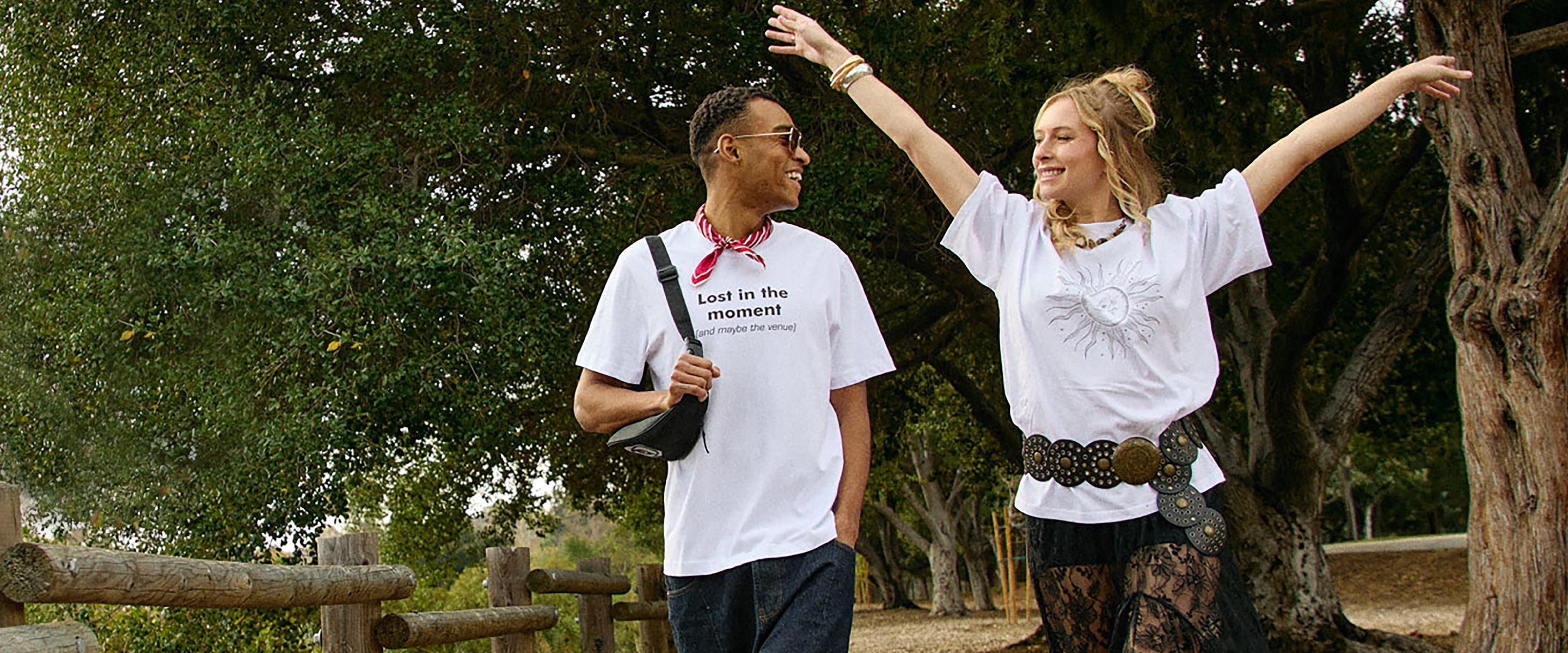 Una joven rubia con una camiseta blanca con estampado y un joven con gafas de sol y camiseta blanca con estampado se miran riendo.
