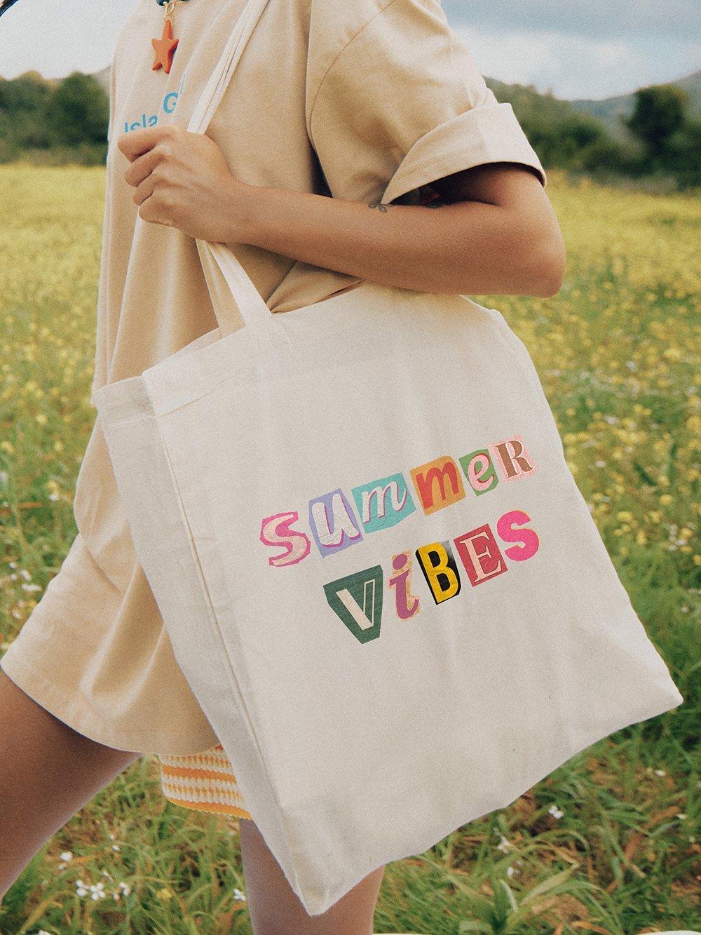Persona sosteniendo una bolsa de tela con la colorida inscripción "Summer Vibes" sobre un prado de flores.