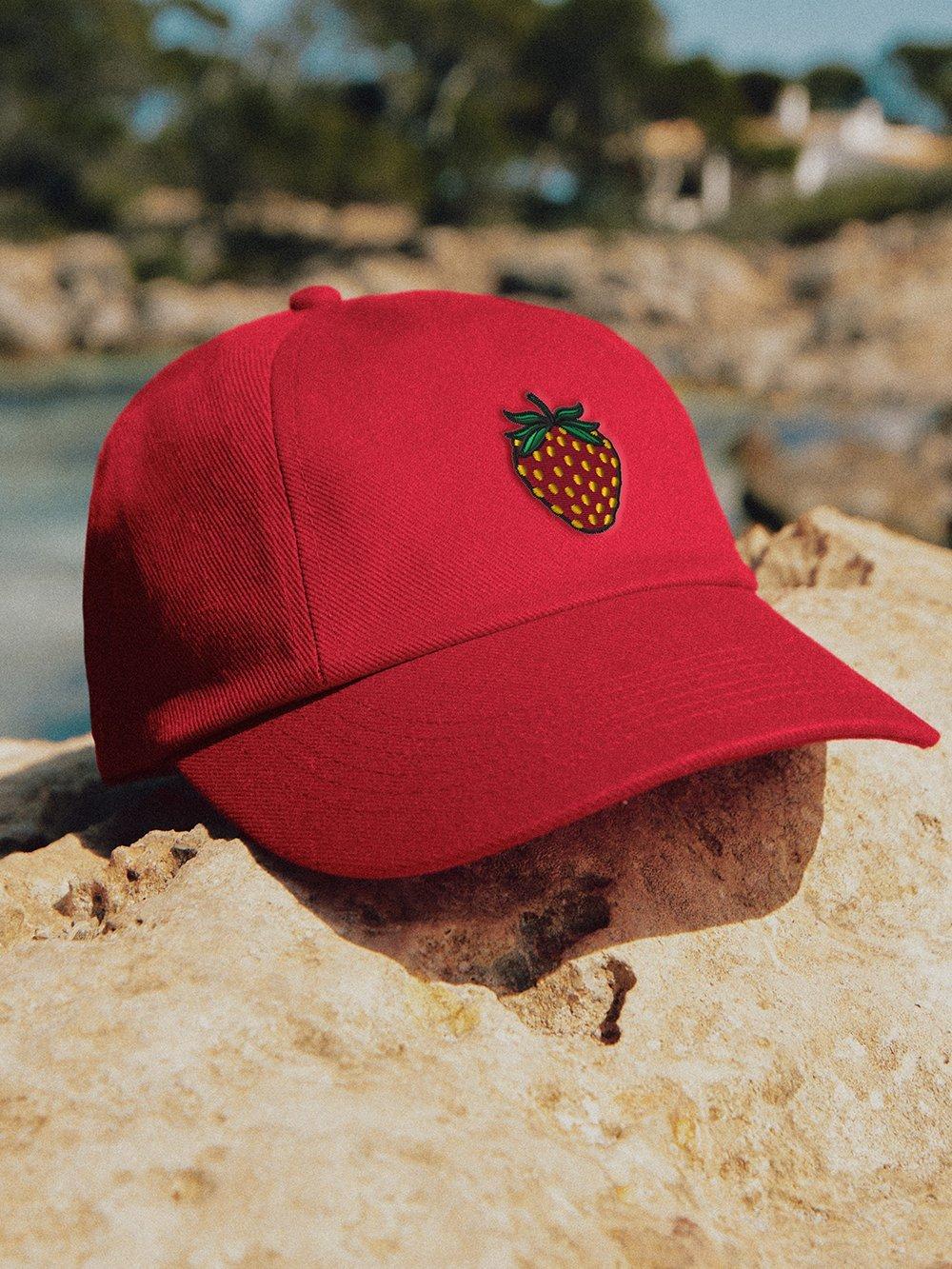 Una gorra roja con una fresa bordada descansa sobre una roca en un entorno veraniego.