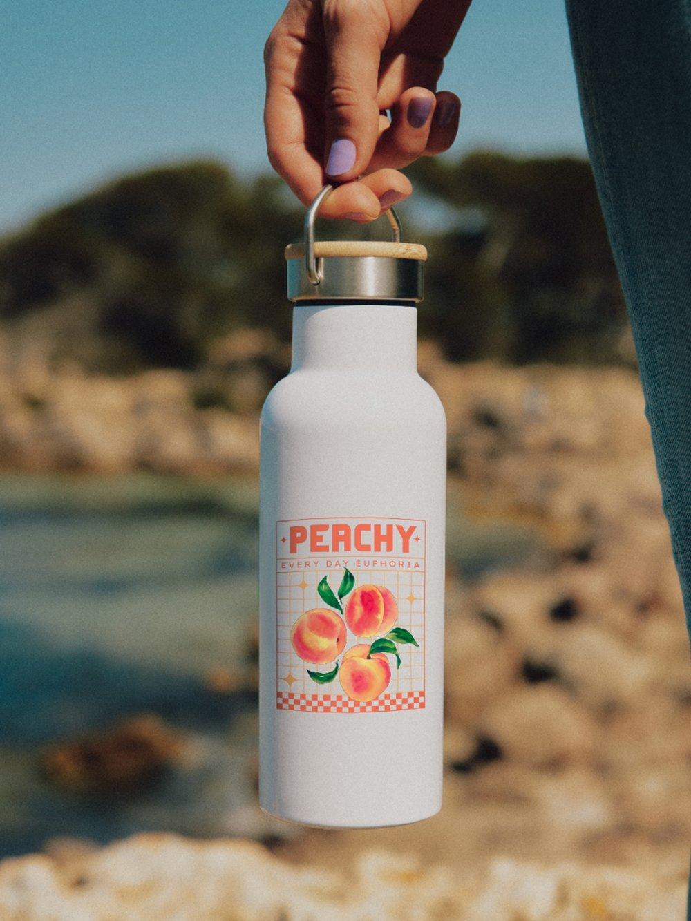 Una mano sostiene una botella blanca para beber con un diseño de melocotón y la palabra "Peachy" (melocotón) en la playa.