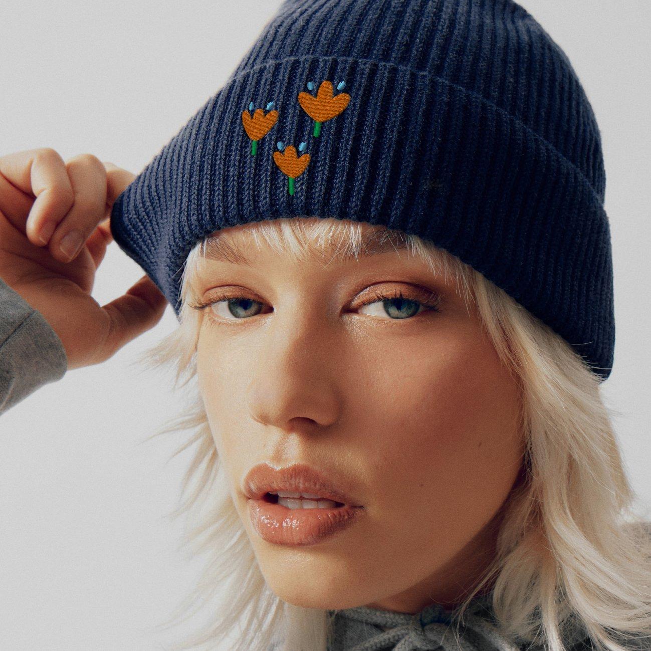 Close-up van een vrouw met blond kapsel en een blauwe gebreide muts met geborduurde oranje bloemen.