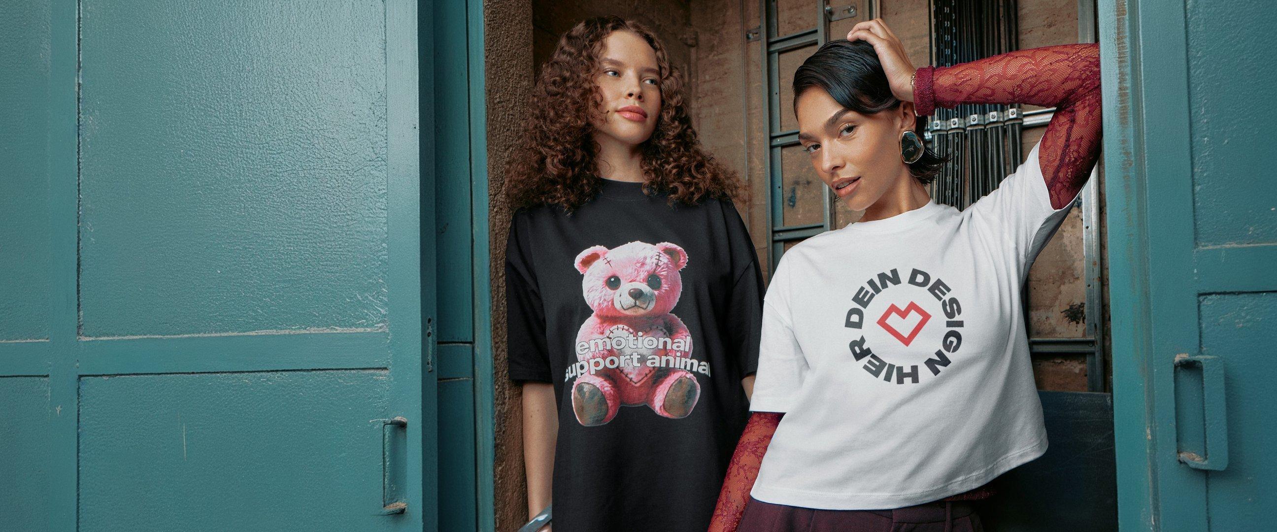 Zwei Frauen posieren in einem industriellen Raum; eine trägt ein schwarzes T-Shirt mit rosa Teddymotiv, die andere ein weißes Shirt mit Kreislogo.